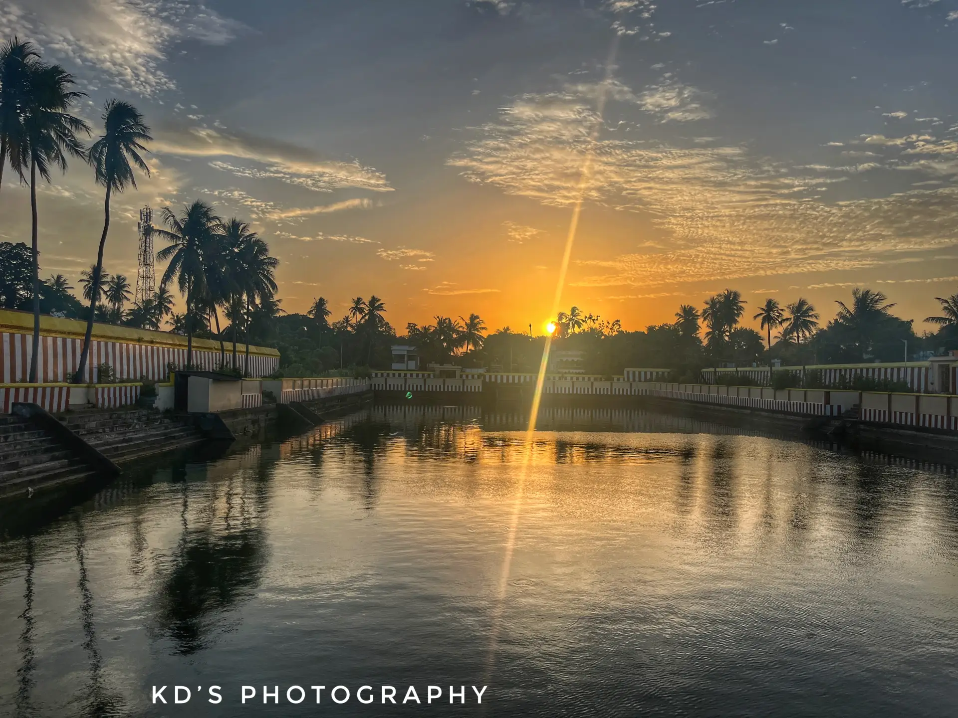 A serene golden sunset over a calm temple tank lined with palm trees and traditional painted walls.