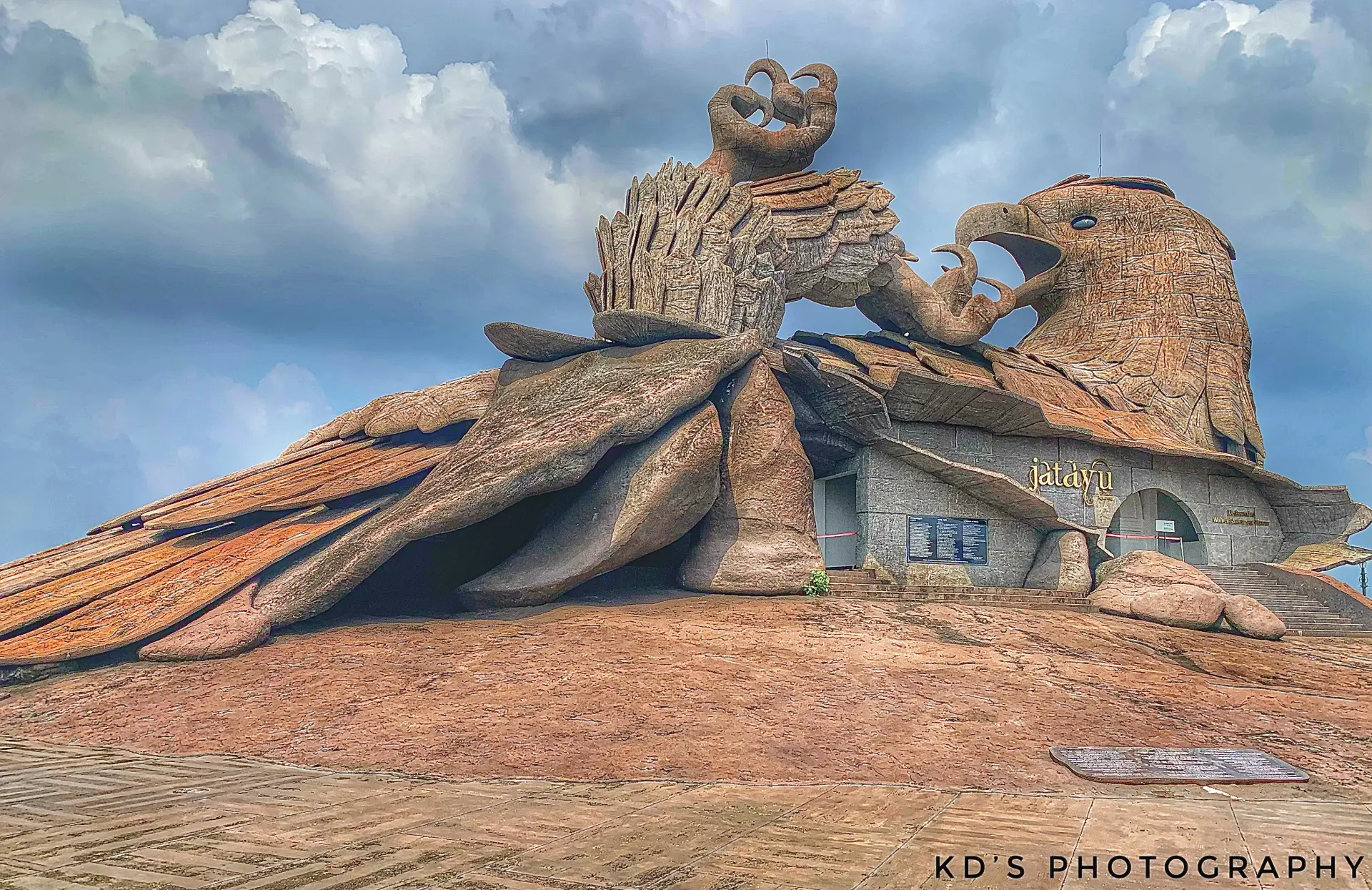 A monumental stone sculpture of a fallen giant eagle resting on a rocky hilltop under a cloudy sky.

